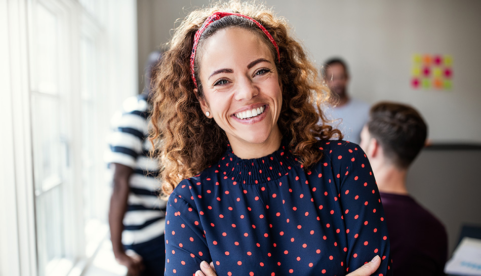 Smiling woman with curly hair standing confidently in a busy office setting.