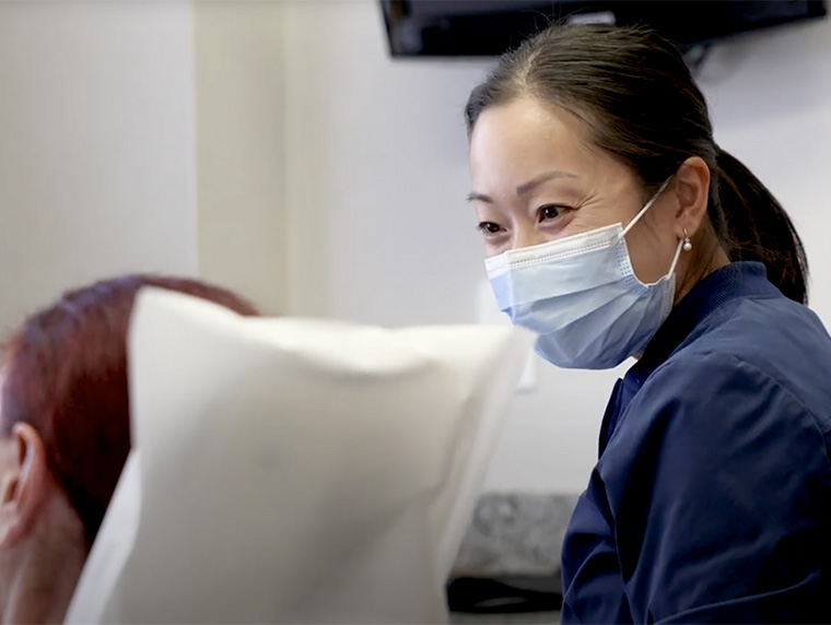 Masked dental professional smiling while speaking with a patient in a treatment room.