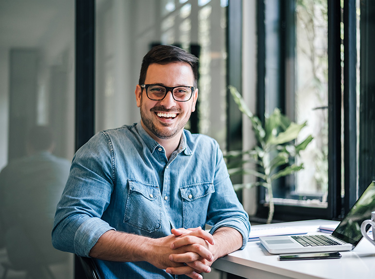 Smiling man with glasses sitting at a desk in a bright modern office.