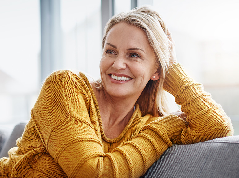 Woman in a mustard sweater smiling while relaxing on a couch at home.