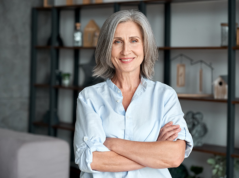 Older woman with gray hair smiling confidently with arms crossed indoors.