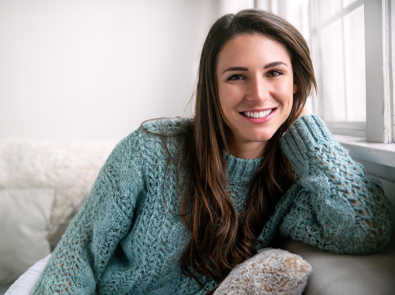 Woman in a knitted sweater smiling while relaxing by a window at home.