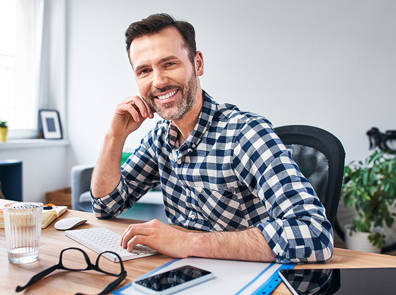Smiling man working at a desk with computer, phone, and office supplies.