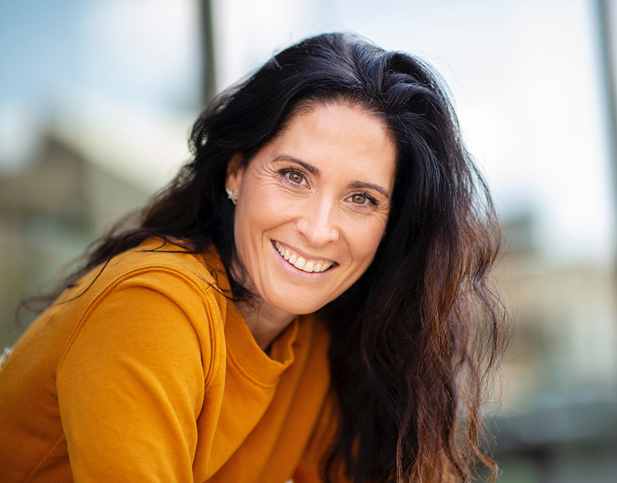 Portrait of a smiling woman with long dark hair wearing a mustard-colored top outdoors.